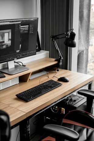 A modern workspace with a large computer monitor displaying 3D modeling software, set on a wooden desk. A keyboard and mouse are placed neatly in front of the monitor. The scene includes a black adjustable desk lamp on the right and a visible computer tower underneath the desk. The workspace is by a window with sheer curtains, allowing natural light to enter.