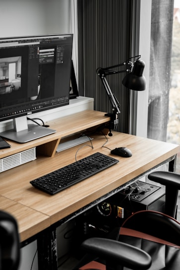A modern workspace with a large computer monitor displaying 3D modeling software, set on a wooden desk. A keyboard and mouse are placed neatly in front of the monitor. The scene includes a black adjustable desk lamp on the right and a visible computer tower underneath the desk. The workspace is by a window with sheer curtains, allowing natural light to enter.