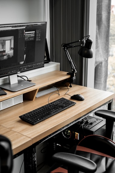A modern workspace with a large computer monitor displaying 3D modeling software, set on a wooden desk. A keyboard and mouse are placed neatly in front of the monitor. The scene includes a black adjustable desk lamp on the right and a visible computer tower underneath the desk. The workspace is by a window with sheer curtains, allowing natural light to enter.