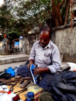 A man is seated on a sidewalk engaged in shoe repair. He is dressed in a striped shirt and checkered pants, intently working on a shoe. Tools and materials, such as scissors, thread, and cans, are scattered around him. The scene is set against an old concrete wall with trees overhead.