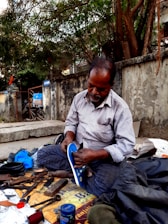 A man is seated on a sidewalk engaged in shoe repair. He is dressed in a striped shirt and checkered pants, intently working on a shoe. Tools and materials, such as scissors, thread, and cans, are scattered around him. The scene is set against an old concrete wall with trees overhead.