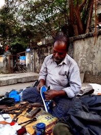 A man is seated on a sidewalk engaged in shoe repair. He is dressed in a striped shirt and checkered pants, intently working on a shoe. Tools and materials, such as scissors, thread, and cans, are scattered around him. The scene is set against an old concrete wall with trees overhead.