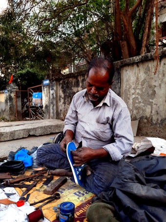 A man is seated on a sidewalk engaged in shoe repair. He is dressed in a striped shirt and checkered pants, intently working on a shoe. Tools and materials, such as scissors, thread, and cans, are scattered around him. The scene is set against an old concrete wall with trees overhead.