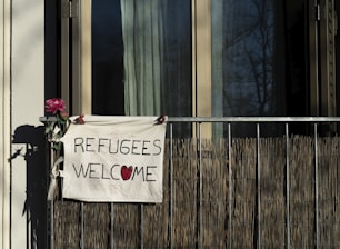 A hand-painted sign hanging on a balcony reads 'REFUGEES WELCOME,' with a red heart symbol replacing the 'O' in 'WELCOME.' A pink flower is attached to the balcony railing beside the sign. The background shows a window partially covered by a curtain.