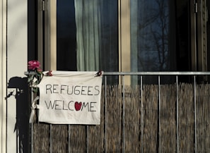 A hand-painted sign hanging on a balcony reads 'REFUGEES WELCOME,' with a red heart symbol replacing the 'O' in 'WELCOME.' A pink flower is attached to the balcony railing beside the sign. The background shows a window partially covered by a curtain.