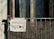 A hand-painted sign hanging on a balcony reads 'REFUGEES WELCOME,' with a red heart symbol replacing the 'O' in 'WELCOME.' A pink flower is attached to the balcony railing beside the sign. The background shows a window partially covered by a curtain.