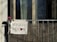 A hand-painted sign hanging on a balcony reads 'REFUGEES WELCOME,' with a red heart symbol replacing the 'O' in 'WELCOME.' A pink flower is attached to the balcony railing beside the sign. The background shows a window partially covered by a curtain.