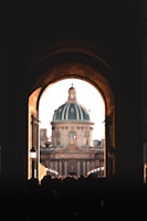 A grand architectural dome is framed by a dark archway, with sunlight illuminating the building's stone exterior. Crowds of people are visible in the foreground, creating a silhouette effect. The French flag is prominently displayed on the building.