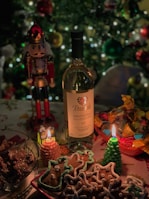 A festive table setting with a bottle of wine, decorated gingerbread cookies, burning candles in the shape of a tree and a house, assorted colorful candies in a bowl, and a nutcracker ornament. The background features a decorated Christmas tree with sparkling lights.