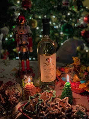 A festive table setting with a bottle of wine, decorated gingerbread cookies, burning candles in the shape of a tree and a house, assorted colorful candies in a bowl, and a nutcracker ornament. The background features a decorated Christmas tree with sparkling lights.