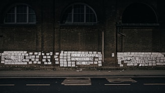 A brick wall is covered with numerous signs and messages, including phrases like 'Say their names' and 'To our murdered sisters'. The messages reference femicides in the UK, emphasizing the number 143 in 2021. The signs are predominantly white with black text, and they are pasted closely together, creating a poignant visual statement.