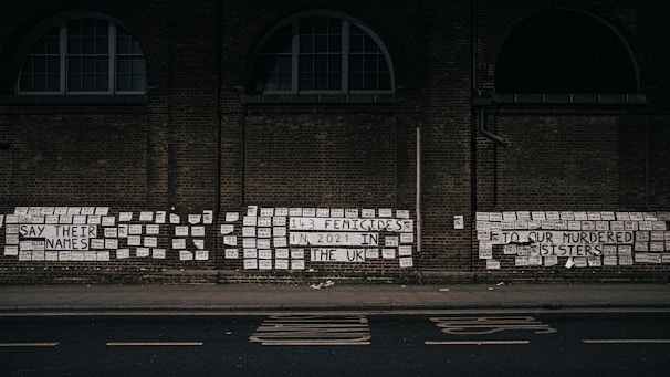 A brick wall is covered with numerous signs and messages, including phrases like 'Say their names' and 'To our murdered sisters'. The messages reference femicides in the UK, emphasizing the number 143 in 2021. The signs are predominantly white with black text, and they are pasted closely together, creating a poignant visual statement.