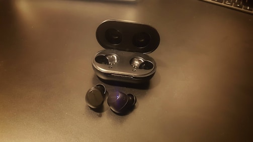 A pair of black wireless earbuds is positioned on a flat surface next to an open charging case. The case is also black with two indentations for storing the earbuds. The lighting creates a subtle reflection on the surface and the earbuds, highlighting their smooth, glossy finish.