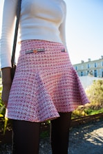 A person is wearing a white long-sleeve top and a textured red and white patterned skirt with decorative buttons. The background shows a clear blue sky and a building with stone architecture, alongside greenery.