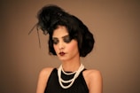 A woman with an elegant hairstyle adorned with feathers wears dark, dramatic makeup. She is dressed in a vintage style, featuring a black sleeveless dress and layered pearl necklaces. The overall look conveys a sense of 1920s fashion and glamour.