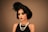 A woman with an elegant hairstyle adorned with feathers wears dark, dramatic makeup. She is dressed in a vintage style, featuring a black sleeveless dress and layered pearl necklaces. The overall look conveys a sense of 1920s fashion and glamour.