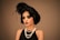 A woman with an elegant hairstyle adorned with feathers wears dark, dramatic makeup. She is dressed in a vintage style, featuring a black sleeveless dress and layered pearl necklaces. The overall look conveys a sense of 1920s fashion and glamour.
