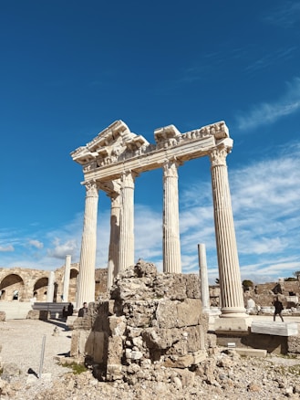 Ancient ruins featuring tall, ornate columns, likely part of a temple structure, stand against a bright blue sky. The foreground includes scattered stone debris and ruins, and a few people can be seen exploring the site.