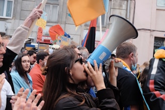 A crowd of people gathered for a protest or demonstration, many holding signs and flags. A woman in the forefront is speaking through a megaphone, while others around her are attentive and engaged.