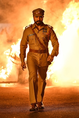 A man in a brown police uniform walks confidently through a scene filled with bright, fiery flames in the background. The setting suggests intensity and action, with the fire creating a dramatic and powerful backdrop.
