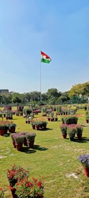 A large, open garden area filled with various potted plants and flowers, arranged neatly on a grassy field. In the background, a tall flagpole with a red, white, and green flag stands prominently. The sky is clear and blue, and the garden is bordered by trees, with a building partially visible to the left.