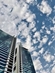 A modern high-rise building with two towers and a glass facade reaching up into a sky filled with scattered clouds.
