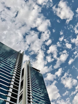 A modern high-rise building with two towers and a glass facade reaching up into a sky filled with scattered clouds.
