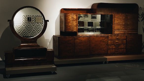 A vintage-style wooden cabinet and dresser with a large circular mirror on the left. The furniture has a rich brown finish with distinct wood grain patterns. The background features a geometric pattern and there are various decorative items placed on and around the furniture.