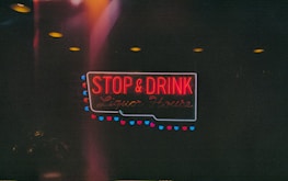 A neon sign with bright red and blue colors displays the words 'STOP & DRINK Liquor House.' The sign is surrounded by small circular lights, creating a moody and dim atmosphere.
