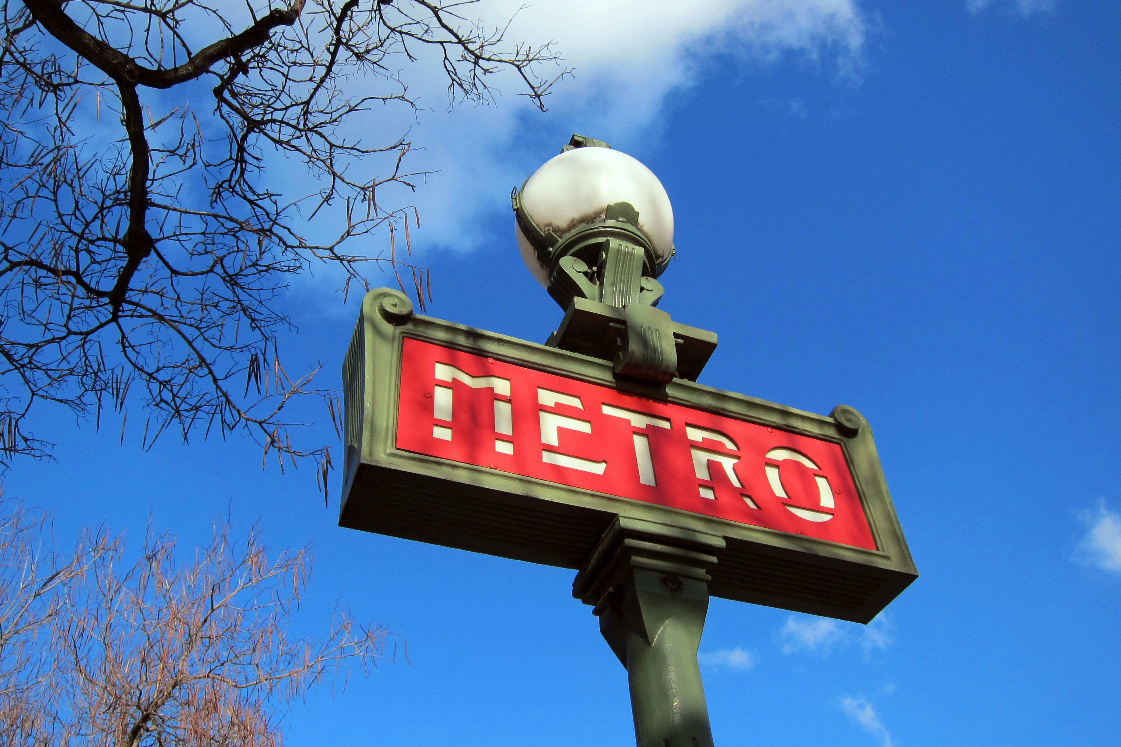a street sign with the word metro on it, This is a metro sign in Paris, France on a sunny day in December. A red sign with white letters reads METRO. It is held up by a green metal pole with a white globe light on the top. Portions of two trees without leaves are visible and the background is a mostly blue sky with a couple white wispy clouds.