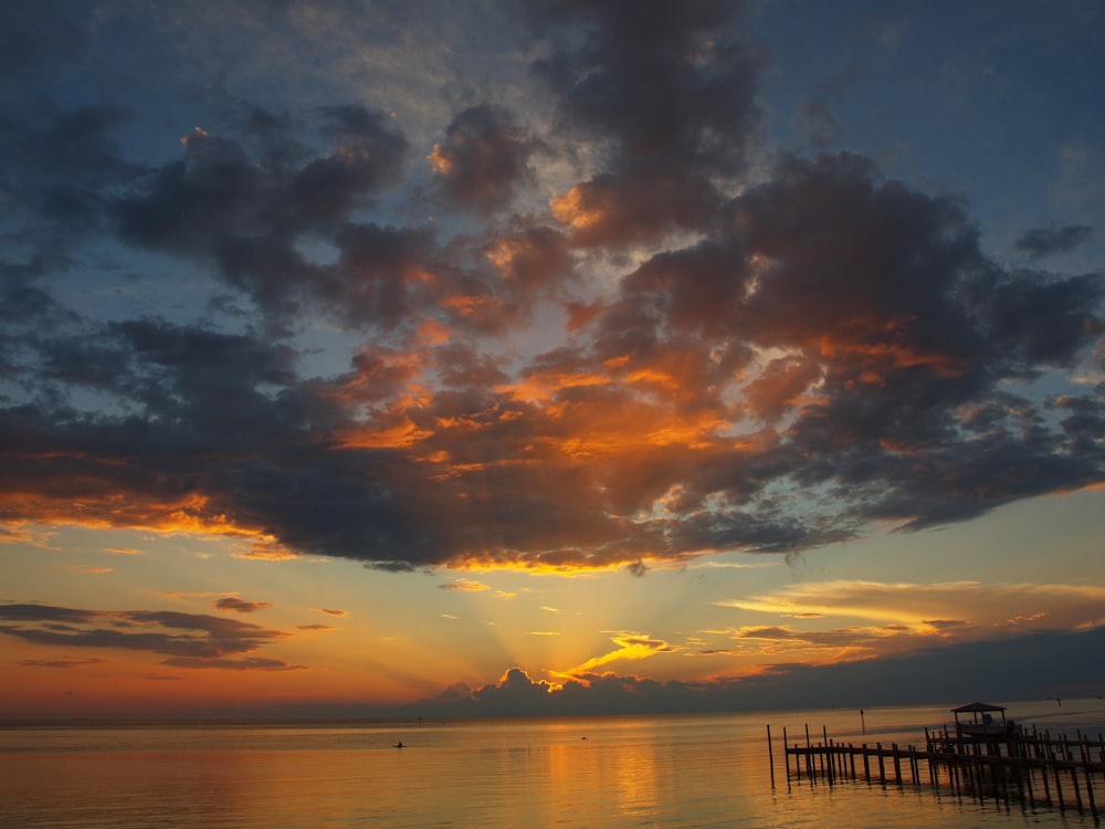 the sun is setting over the ocean with a pier in the foreground