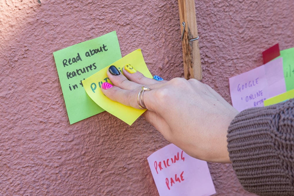 a mão de uma mulher em uma nota de post it anexada a uma parede rosa