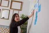 A woman with dark hair gestures towards a series of sticky notes on a wall. The room has a creative and professional ambiance, with empty picture frames hanging on another wall and a tall plant nearby. The sticky notes are in various colors, predominantly blue and yellow, suggesting brainstorming or planning.