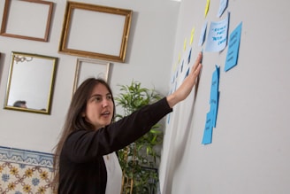 A woman with dark hair gestures towards a series of sticky notes on a wall. The room has a creative and professional ambiance, with empty picture frames hanging on another wall and a tall plant nearby. The sticky notes are in various colors, predominantly blue and yellow, suggesting brainstorming or planning.