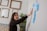 A woman with dark hair gestures towards a series of sticky notes on a wall. The room has a creative and professional ambiance, with empty picture frames hanging on another wall and a tall plant nearby. The sticky notes are in various colors, predominantly blue and yellow, suggesting brainstorming or planning.