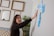 A woman with dark hair gestures towards a series of sticky notes on a wall. The room has a creative and professional ambiance, with empty picture frames hanging on another wall and a tall plant nearby. The sticky notes are in various colors, predominantly blue and yellow, suggesting brainstorming or planning.
