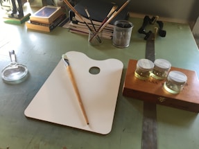 A workspace featuring an artist's palette with a paintbrush resting on it, three small jars of liquid on a wooden box, and a magnifying glass. Books and notebooks are stacked in the background, along with a pen holder containing various writing instruments.