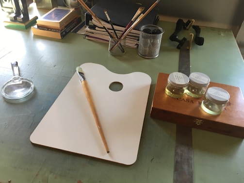 A workspace featuring an artist's palette with a paintbrush resting on it, three small jars of liquid on a wooden box, and a magnifying glass. Books and notebooks are stacked in the background, along with a pen holder containing various writing instruments.