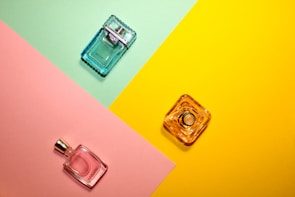 Three perfume bottles are arranged on a colorful background divided into sections of mint green, pink, and yellow. The bottles are of different shapes and colors: one is a blue square bottle positioned on the green section, a pink bottle on the pink section, and a clear yellow bottle on the yellow section.