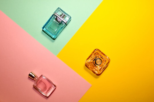 Three perfume bottles are arranged on a colorful background divided into sections of mint green, pink, and yellow. The bottles are of different shapes and colors: one is a blue square bottle positioned on the green section, a pink bottle on the pink section, and a clear yellow bottle on the yellow section.