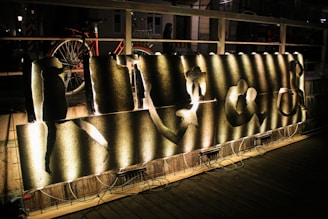 A piece of art is displayed on a wooden deck, illuminated by arranged lighting from below. The artwork consists of several metallic panels with abstract cutouts, each lit to create various shadows and patterns on the surface. In the background, a bicycle is parked against a wooden railing, and a person is partially visible. The setting appears to be outdoors at night.