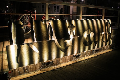 A piece of art is displayed on a wooden deck, illuminated by arranged lighting from below. The artwork consists of several metallic panels with abstract cutouts, each lit to create various shadows and patterns on the surface. In the background, a bicycle is parked against a wooden railing, and a person is partially visible. The setting appears to be outdoors at night.