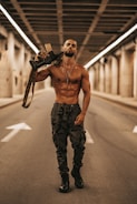 A muscular man is walking down an empty street under a bridge or tunnel, carrying a rifle over his shoulder. He is shirtless, wearing camouflage pants and black boots, with a pendant necklace. The setting is urban and has an industrial feel, with concrete structures and directional arrows on the road.