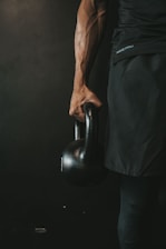A person is holding a kettlebell in their right hand. The image focuses on the muscular arm and the handle of the kettlebell, with a dark and moody background. The person is wearing dark athletic apparel.