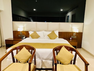 A modern hotel room with a large bed featuring white linens and mustard yellow accent pillows. Two matching wooden chairs with yellow striped cushions are positioned at the foot of the bed. The room includes bedside tables with lamps providing soft lighting and a dark, reflective wall behind the bed adding depth.