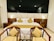 A modern hotel room with a large bed featuring white linens and mustard yellow accent pillows. Two matching wooden chairs with yellow striped cushions are positioned at the foot of the bed. The room includes bedside tables with lamps providing soft lighting and a dark, reflective wall behind the bed adding depth.