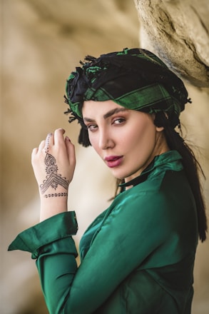 A woman poses with a confident expression, wearing a green shirt and a matching headscarf. Her hand is delicately positioned near her face, showcasing intricate henna designs on her skin. The background is a soft-focus natural setting, creating a serene and artistic ambiance.