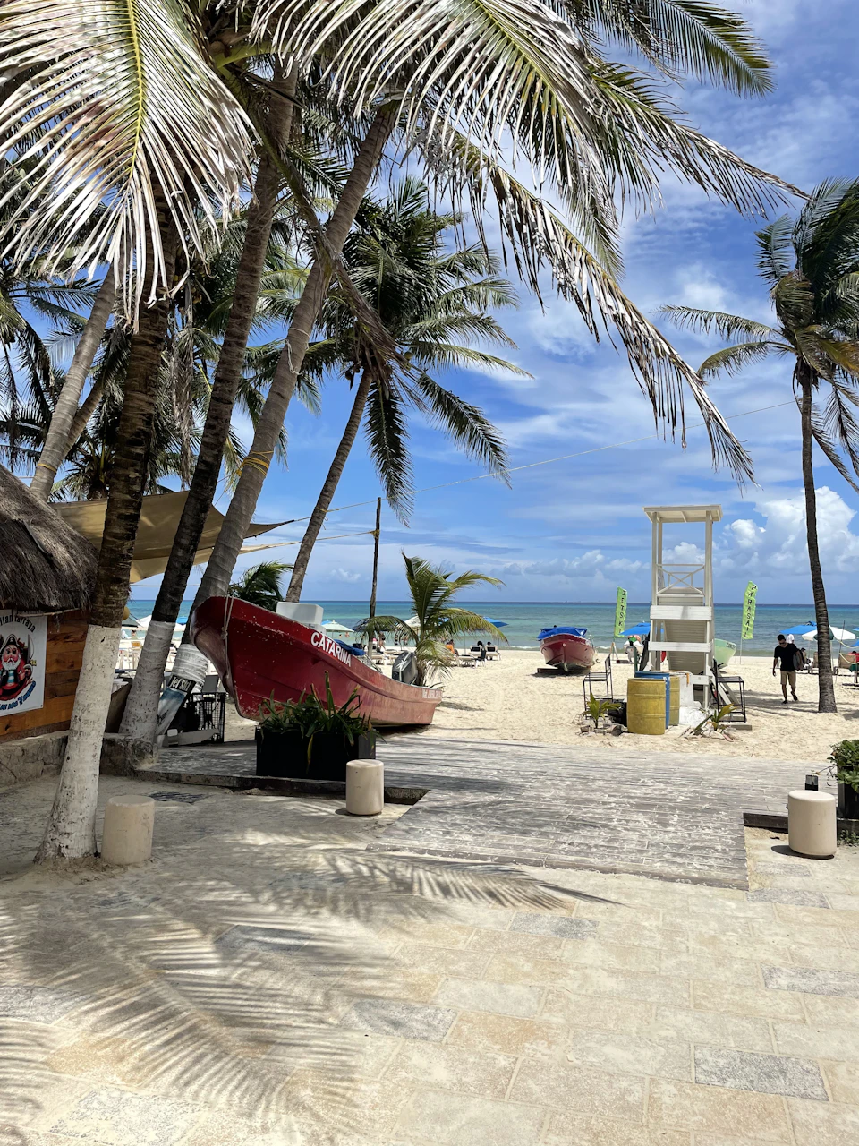 The image depicts a serene tropical beach scene with palm trees swaying gently against a clear blue sky. The sandy beach is dotted with several people enjoying their day, some lounging on chairs and others walking along the shoreline. A red boat is moored near the water's edge, adding to the picturesque setting. In the background, there are buildings visible, suggesting that this beach may be located in a resort area or a popular tourist destination.

The image also contains several objects such as a chair and a potted plant placed on the sandy beach, contributing to the overall ambiance of relaxation and leisure.