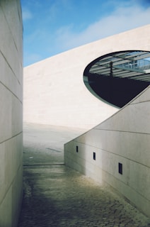 A modern architectural scene featuring clean, sturdy walls made of large stone slabs with smooth textures. The area is open to the sky, which is clear with a few clouds. An oval-shaped cut-out in one of the walls reveals a grid-like structure in the background, possibly part of a building's interior or roof.