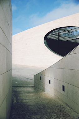 A modern architectural scene featuring clean, sturdy walls made of large stone slabs with smooth textures. The area is open to the sky, which is clear with a few clouds. An oval-shaped cut-out in one of the walls reveals a grid-like structure in the background, possibly part of a building's interior or roof.